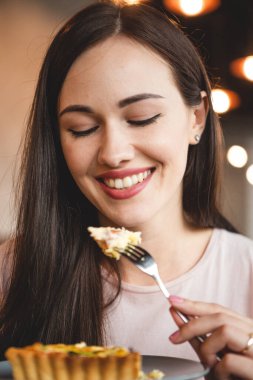 Enfes bir gülümsemesi ve güzel gözleri olan bir kadın, samimi bir kafede arkadaşıyla kahvaltı yapıyor. Çatal yer, kek yer ve masada bir sandalyede oturup çay içer.