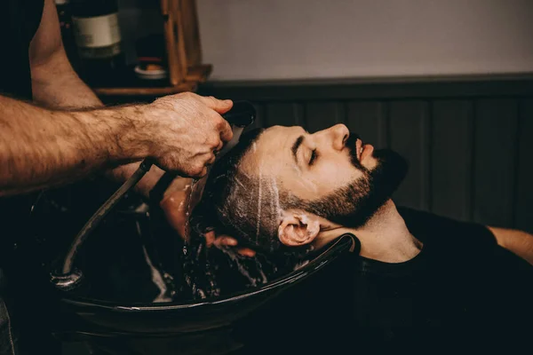 Handsome man with a beard in the barbershop. His head lies in the white sink and a barber washes it with a black faucet. Guy looks up. Close up.