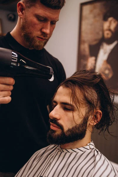 Barbershop. Close-up of man haircut, master does the hair styling in barber shop. Strong muscular bearded barber cuts long black hair of the client