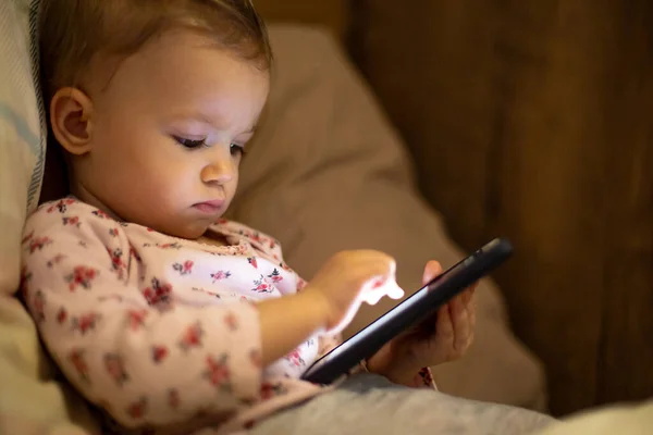 Işıklar kapalı bir odada, elinde akıllı bir telefonla, yatakta uzanmış oyun oynayan ve çizgi film izleyen küçük bir kız. Aletlere bebek bağımlılığı