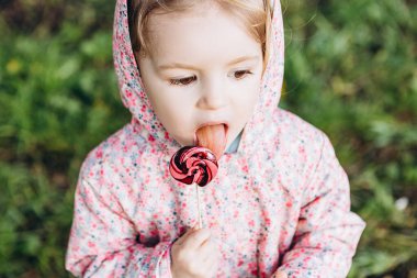3 yaşındaki küçük bir kız yağmurlu bir yaz gününde parkta yürürken renkli bir girdap şekeri yalıyor.