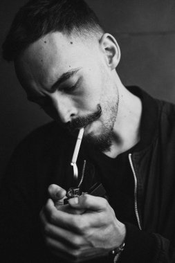 Genç, vahşi sakallı, şık bıyıklı bir adam sigarasını yakıyor ve sigara içiyor. Siyah beyaz fotoğraf