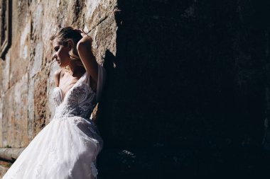 Uzun beyaz bir elbise giymiş genç, seksi bir gelin büyük bir taş duvarın arka planında fotoğraf için poz veriyor. Mesajınız için kopya alanı.