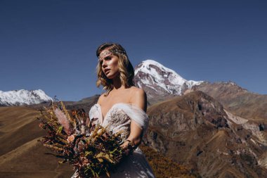 Gelinlik giymiş, boho tarzı genç ve çekici bir kadın. Dağlarda fotoğraf çektirmek için poz veriyor. Makyaj, saç stili ve düğün çiçekçisi.