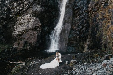 Ses efekti, seçici odak: İnanılmaz derecede aşık gelinlerin kucaklaşması, öpüşmesi ve poz vermesi inanılmaz kayalık arka planda büyük bir şelale ile birlikte.