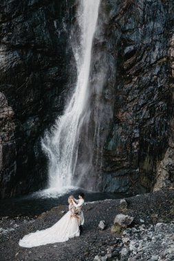 Ses efekti, seçici odak: İnanılmaz derecede aşık gelinlerin kucaklaşması, öpüşmesi ve poz vermesi inanılmaz kayalık arka planda büyük bir şelale ile birlikte.
