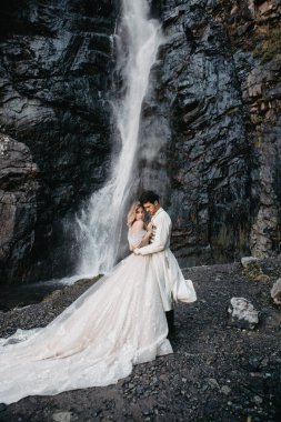 Ses efekti, seçici odak: İnanılmaz derecede aşık gelinlerin kucaklaşması, öpüşmesi ve poz vermesi inanılmaz kayalık arka planda büyük bir şelale ile birlikte.