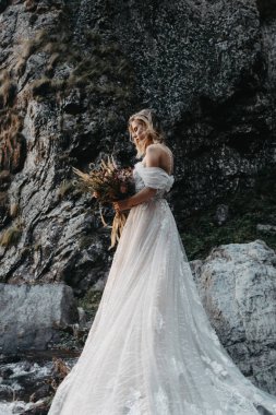 Gürültü efekti, seçici odak: dağ taşlarının arka planında şık uzun gelinlik giymiş çekici bir kız, düğün salonu için promosyon fotoğrafları için poz veriyor.
