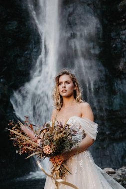 Dağ taşlarının arkasında şık, uzun bir gelinlik giymiş çekici bir kız. Düğün salonunun promosyon fotoğrafları için poz veriyor.