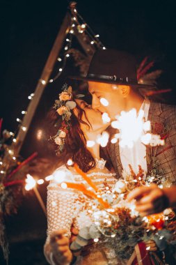 Şık bir çift gelin, gelinlik giymiş güzel bir kadın ve fötr şapkalı genç bir adam. Doğada romantik bir düğün töreni. Elektrikli çelengin ışıkları düğün partisini aydınlatıyor..