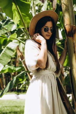 Uzun keten bir elbise giymiş şık, çekici bir kadın. Yazın arka planda palmiye ağaçları ve parlak güneşli denizde çekilmiş bir şapkası var.