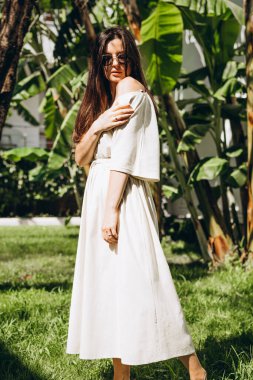 Uzun keten bir elbise giymiş şık, çekici bir kadın. Yazın arka planda palmiye ağaçları ve parlak güneşli denizde çekilmiş bir şapkası var.