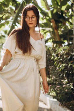 Uzun keten bir elbise giymiş şık, çekici bir kadın. Yazın arka planda palmiye ağaçları ve parlak güneşli denizde çekilmiş bir şapkası var.