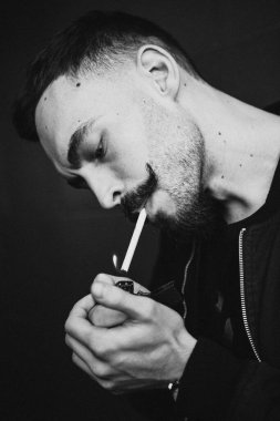 Genç, vahşi sakallı, şık bıyıklı bir adam sigarasını yakıyor ve sigara içiyor. Siyah beyaz fotoğraf