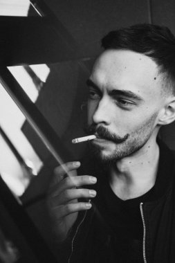 Genç, vahşi sakallı, şık bıyıklı bir adam sigarasını yakıyor ve sigara içiyor. Siyah beyaz fotoğraf