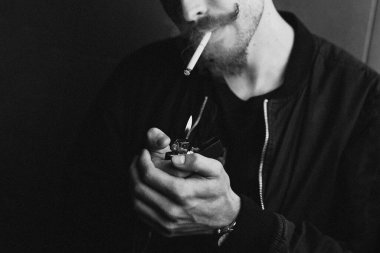 Genç, vahşi sakallı, şık bıyıklı bir adam sigarasını yakıyor ve sigara içiyor. Siyah beyaz fotoğraf