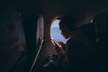 Kadın ilk kez uçuyor ve pencerenin yanındaki sandalyede otururken telefondaki fotoğrafa bakıyor.