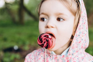 3 yaşındaki küçük bir kız yağmurlu bir yaz gününde parkta yürürken renkli bir girdap şekeri yalıyor.
