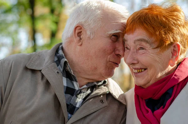 A couple of elderly people. A man with gray hair and a woman with gray hair colored in red color sitting next to each other in the park.