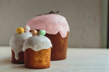 Üç küçük Paskalya pastası, beyaz ahşap bir masanın üzerinde jöleli ve dekoratif renkli çikolata yumurtalarıyla süslenmiş. Metnin için yer