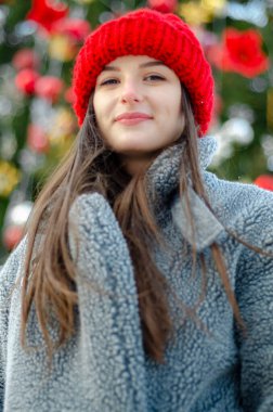 Genç, güzel model esmer, kışın, şehir parkının ortasında, kırmızı örgü bir şapka ve sıcak gri bir ceket ve eldiven giymiş bir Noel ağacının yanında poz veriyor.