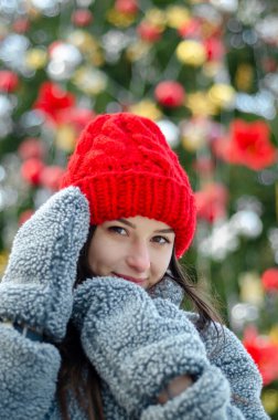 Genç, güzel model esmer, kışın, şehir parkının ortasında, kırmızı örgü bir şapka ve sıcak gri bir ceket ve eldiven giymiş bir Noel ağacının yanında poz veriyor.