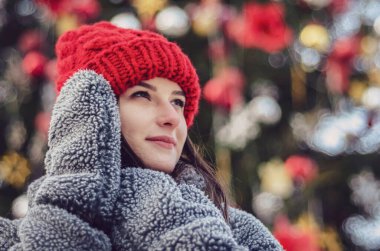 Genç, güzel model esmer, kışın, şehir parkının ortasında, kırmızı örgü bir şapka ve sıcak gri bir ceket ve eldiven giymiş bir Noel ağacının yanında poz veriyor.