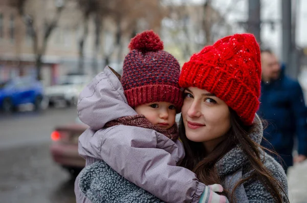 mother with baby on her hands in winter in the middle of a snow-covered winter street in the city, the child is asleep and shrugged face to mother. Mother in gloves and red hats