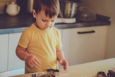 Küçük bir çocuk mutfakta hamur ve un dolu bir masanın üzerinde makarna, kurabiye, ekmek, zencefilli ekmek gibi çeşitli yemekler pişiriyor. Mutfakta aile eğlencesi kavramı, çocuk yemek hazırlıyor..