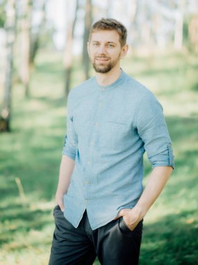 Sarı saçlı, mavi gömlekli şık sakallı adam parkta fotoğraf çektirmek için poz veriyor..