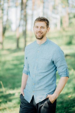 Sarı saçlı, mavi gömlekli şık sakallı adam parkta fotoğraf çektirmek için poz veriyor..