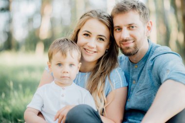 Mutlu ebeveynler ormanda yeşil çimlerin üzerinde yatan oğullarına sarılıyorlar. Genç ve mutlu bir aile kavramı. Ebeveynler çocukla birlikte gülümser. İdeal aile