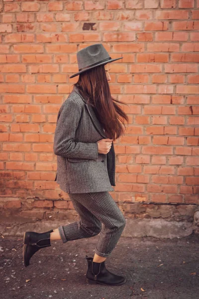 a stylish woman in a classic women\'s suit consisting of trousers and a jacket in a hood in the style of a gangster, posing for a photo against the background of a brick wall