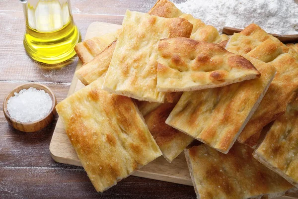 Focaccia bread, flat bread, with chopping board, olive oil, flour and salt on wooden floured background. Roman white pizza, a thin and fragrant focaccia, closeup.