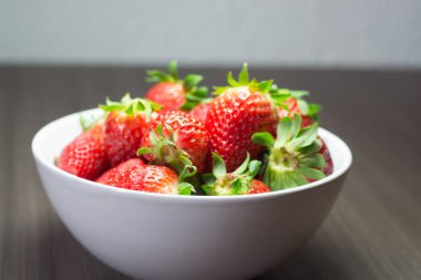 Taze organik çileklerin fotoğraf çekimi. Ayrıca gri ahşap dokular ve beyaz bir kase içeriyor. Parlak renklerin birleşimi öne çıkıyor.