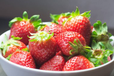 Taze organik çileklerin fotoğraf çekimi. Ayrıca gri ahşap dokular ve beyaz bir kase içeriyor. Parlak renklerin birleşimi öne çıkıyor.