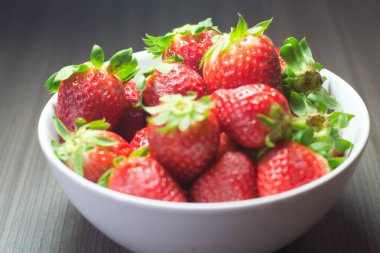 Taze organik çileklerin fotoğraf çekimi. Ayrıca gri ahşap dokular ve beyaz bir kase içeriyor. Parlak renklerin birleşimi öne çıkıyor.