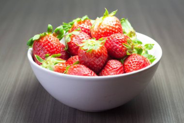 Taze organik çileklerin fotoğraf çekimi. Ayrıca gri ahşap dokular ve beyaz bir kase içeriyor. Parlak renklerin birleşimi öne çıkıyor.