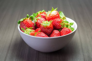 Taze organik çileklerin fotoğraf çekimi. Ayrıca gri ahşap dokular ve beyaz bir kase içeriyor. Parlak renklerin birleşimi öne çıkıyor.