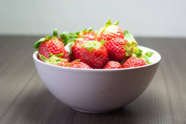 Taze organik çileklerin fotoğraf çekimi. Ayrıca gri ahşap dokular ve beyaz bir kase içeriyor. Parlak renklerin birleşimi öne çıkıyor.