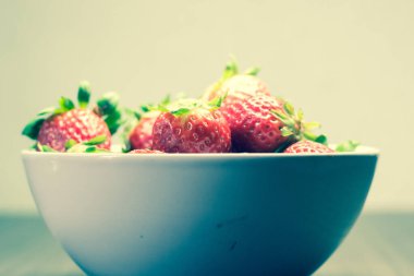 Taze organik çileklerin fotoğraf çekimi. Ayrıca gri ahşap dokular ve beyaz bir kase içeriyor. Parlak renklerin birleşimi öne çıkıyor.