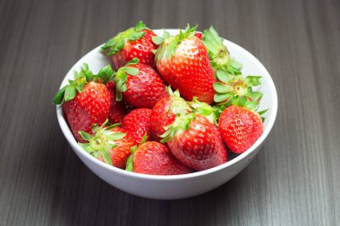 Taze organik çileklerin fotoğraf çekimi. Ayrıca gri ahşap dokular ve beyaz bir kase içeriyor. Parlak renklerin birleşimi öne çıkıyor.