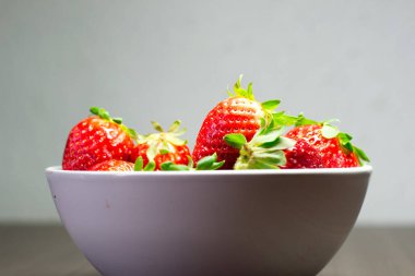 Taze organik çileklerin fotoğraf çekimi. Ayrıca gri ahşap dokular ve beyaz bir kase içeriyor. Parlak renklerin birleşimi öne çıkıyor.
