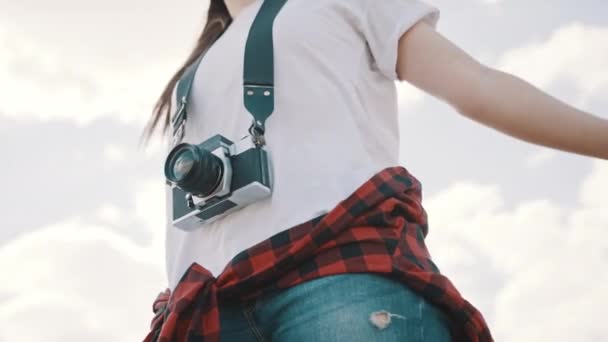 Молодая женщина с винтажной камерой и протянутыми руками наслаждается бризом. Снимок под низким углом с голубым небом на фоне облаков — стоковое видео