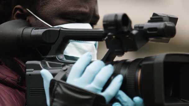 Camarógrafo africano con máscara médica sosteniendo la cámara con los guantes de látex. Informes sobre pandemia de coronavirus . — Vídeos de Stock