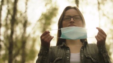 Gözlüklü genç bir kadın yüzüne tıbbi maske takıyor. Güneş ışınları arkadaki dallardan geliyor. Ağır çekim portre.