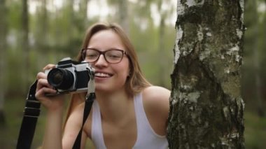 Ağacın arkasında gizlenen beyaz bir kadın, eski model bir kamerayla. Doğayı fotoğraflayan mutlu kız.