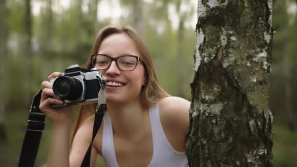 Młoda biała kobieta chowa się za drzewem z zabytkową kamerą. Szczęśliwa dziewczyna fotografowania przyrody — Wideo stockowe