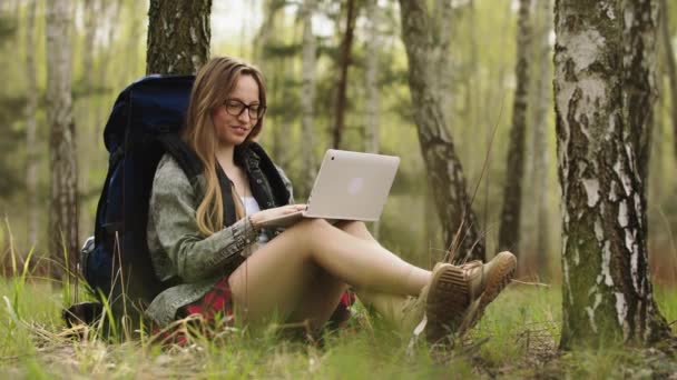 Νεαρή γυναίκα με μεγάλο χαμόγελο, σακίδιο πλάτης και γυαλιά κάθεται στο πάρκο και χρησιμοποιεί φορητό υπολογιστή. Έννοια ταξιδιών και blogging — Αρχείο Βίντεο