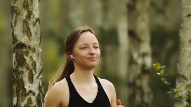 Gesunder Lebensstil. Junge kaukasische Frau mit kabellosen Kopfhörern, die durch den Wald läuft. Porträt — Stockvideo